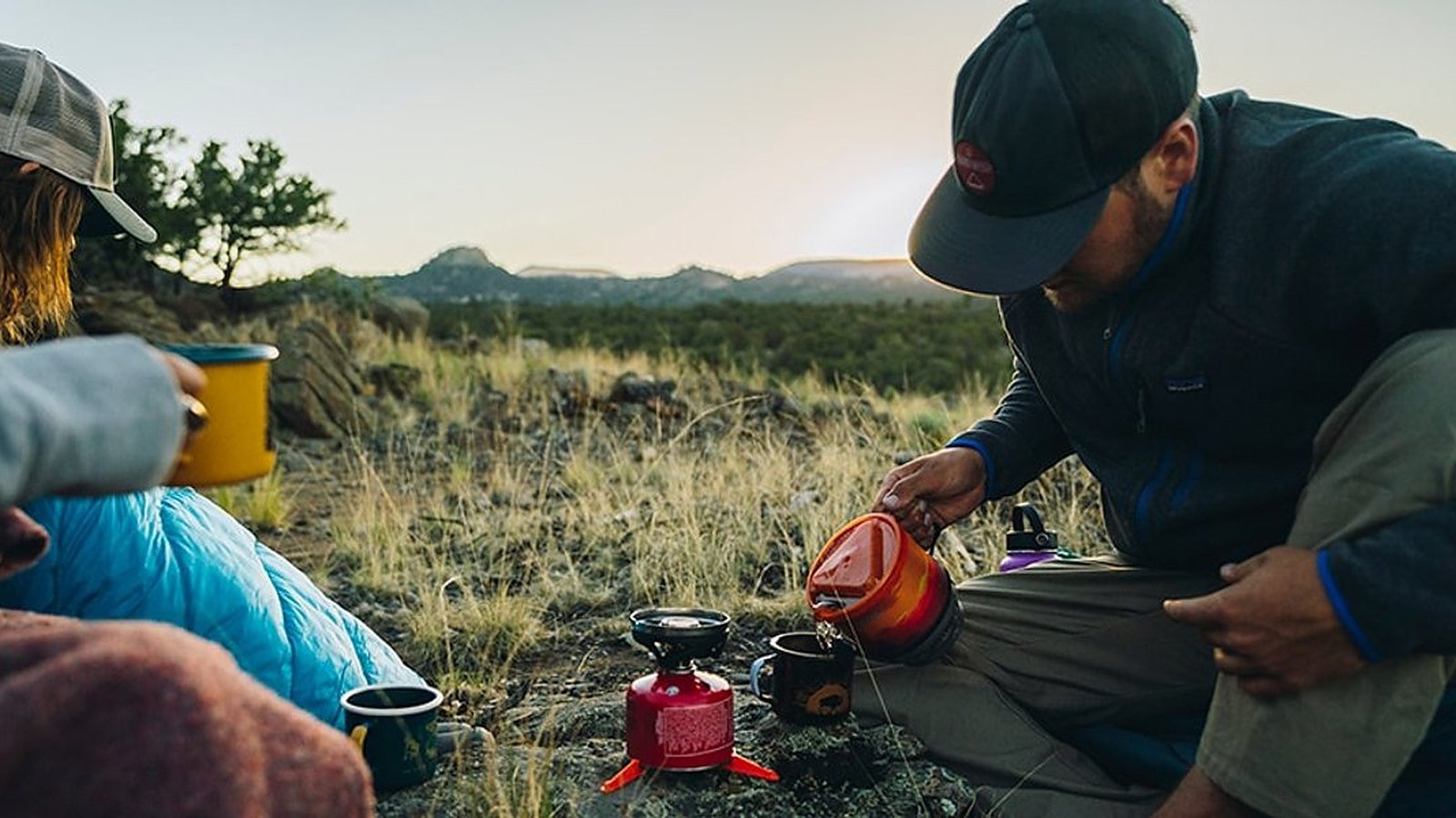 Two campers sharing coffee at sunrise, one pouring hot water from a camp stove pot into a mug