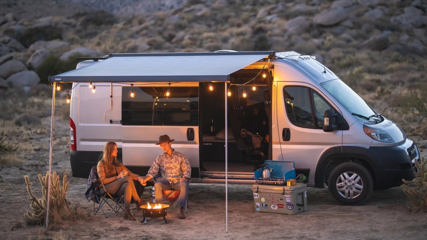 A couple sitting by a small fire pit next to a camper van with an awning and string lights at dusk in the desert, a camp stove and kettle set up on a cooler